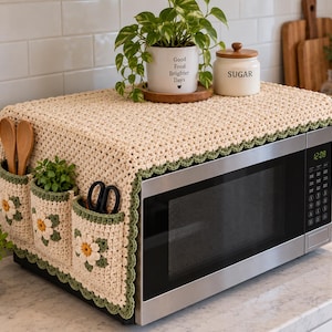 May include: A cream-colored crochet microwave cover featuring a scalloped sage green border and three front pockets adorned with white and yellow daisy motifs. The cover is draped over a stainless steel microwave with a digital display showing 12:08.
