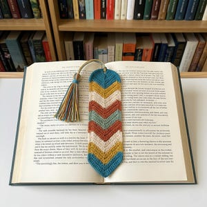 Puede incluir: Marcapáginas de ganchillo con un diseño de chevrón en colores turquesa, amarillo, naranja y crema. El marcapáginas tiene una borla y está colocado en un libro abierto. Un artículo decorativo para lectores.