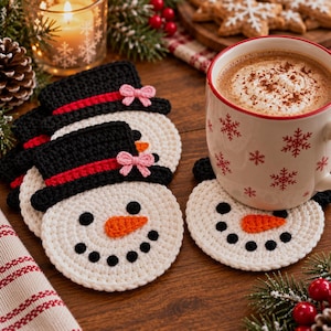 May include: A set of round, white crocheted snowman coasters featuring black top hats with red bands and small pink bows. Each snowman face has black circular eyes, a stitched orange carrot nose, and a black dotted smile. One coaster sits under a white mug.
