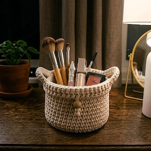 May include: A cream-coloured, hand-crocheted basket contains makeup brushes, tubes, and a compact. The basket has two handles and a wooden button closure. The makeup brushes have wooden handles and brown bristles. The basket sits on a dark wooden surface.