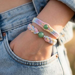 May include: Two pastel-colored bracelets. One is braided with a large green bead and silver accents. The other is crocheted with a green bead. The bracelets are on a wrist, with a denim jacket and jeans in the background.