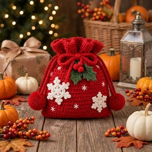 Pode incluir: Uma bolsa de crochê vermelha com cordão, decorada com aplicações de flocos de neve brancos, folhas de azevinho verdes e bagas vermelhas, com dois pompons vermelhos nas extremidades dos cordões. A bolsa tem uma textura artesanal.