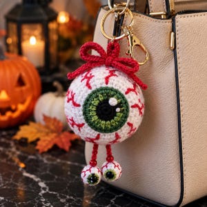 May include: A crocheted eyeball keychain featuring a large white sphere with red veins, a green iris, and a black pupil. A red crocheted bow sits on top, and two smaller dangling eyeballs hang from the bottom, all attached to a gold metal keyring.