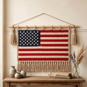 Könnte beinhalten: Gehäkelte Wanddekoration mit der amerikanischen Flagge. Die Flagge hat rote und weiße Streifen, ein blaues Feld mit weißen Sternen und eine Fransenbordüre. Sie hängt an einer Holzstange mit Quasten. Ein Symbol der Vereinigten Staaten.