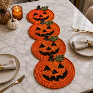 Può includere: Un runner da tavola all'uncinetto con quattro motivi circolari a forma di zucca arancione, caratterizzati da volti di jack-o'-lantern cuciti in nero e piccole foglie verdi. Le zucche sono collegate in una fila verticale decorativa.