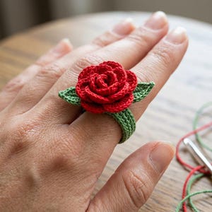 May include: A handmade crochet ring featuring a vibrant red rose with green leaves. The ring band is green, and the rose is the focal point. The ring is displayed on a hand, showcasing the intricate details of the floral design.