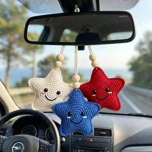 May include: Crocheted car charm featuring three smiling stars in red, white, and blue. The stars are suspended from a string with wooden beads, hanging from a car's rearview mirror. The charm adds a decorative touch to the vehicle's interior.