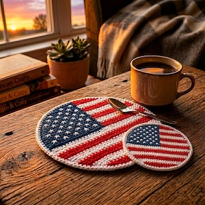 May include: A crocheted coaster set featuring the American flag design. The set includes a large round coaster and a smaller matching coaster. The coasters are red, white, and blue, with a white border. A mug of coffee and a spoon are also on the wooden table.