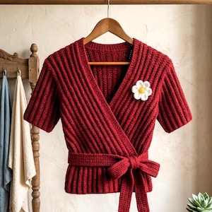 May include: A red knitted wrap top with short sleeves and a matching tie belt. The top features a ribbed texture and a white and yellow flower pin. The garment is displayed on a wooden hanger.