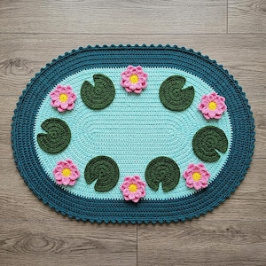 May include: A handmade, oval-shaped crochet rug featuring a lily pad design. The rug has a light blue center, a dark teal border, and is adorned with green lily pads and pink and yellow flowers. The rug is placed on a wooden surface.
