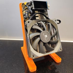 Peut inclure: Un composant informatique avec un ventilateur gris et un centre noir, monté sur une carte de circuit imprimé noire. Il est soutenu par un support orange. Un petit écran noir est visible en haut.
