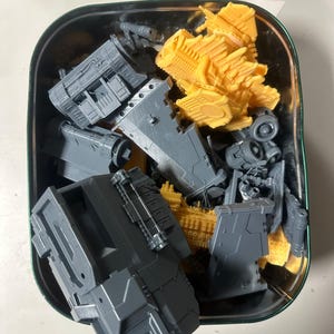 Può includere: Una collezione di parti di modellismo in plastica grigia e gialla non verniciate, conservate all'interno di una scatola di metallo rettangolare. I componenti presentano dettagli meccanici complessi, inclusi pannelli, prese d'aria e strutture.