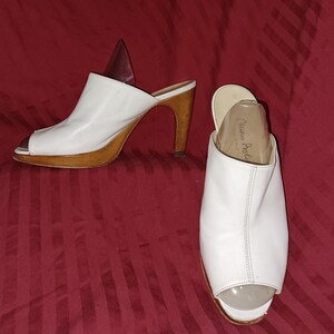 Könnte beinhalten: Weiße Leder-Peeptoe-Mules mit Holzabsätzen und Plateaus. Die Schuhe haben ein klassisches Design mit hohem Absatz und Slip-on-Stil. Die Absätze sind etwa 7,6 cm hoch.
