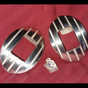 May include: Two oval-shaped, silver-toned belt buckles with a diagonal striped design and a rectangular opening in the center. The buckles are displayed on a red background. A small tag is attached.