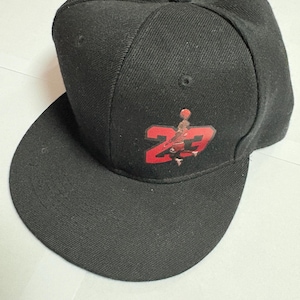 Puede incluir: Gorra de béisbol negra con visera plana. La gorra presenta un logotipo rojo y negro con el número 23. La gorra está hecha de una tela texturizada y tiene una corona estructurada.
