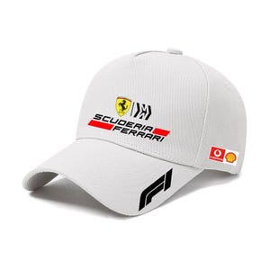 Puede incluir: Gorra de béisbol blanca con el logotipo de Scuderia Ferrari en negro, rojo y amarillo. La gorra tiene una visera curva y un logotipo de Fórmula 1 en el lateral. También son visibles los logotipos de Vodafone y Shell.