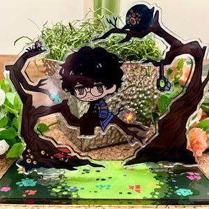以下が含まれることがあります： メガネをかけた漫画のキャラクターが、様式化された木のデザインの中に配置された透明なアクリルディスプレイ。ベースは、カラフルなアクセントのある緑の風景です。小さな生き物と光るオブジェクトも存在します。