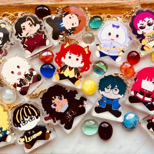 Könnte beinhalten: Eine Sammlung von durchsichtigen Acryl-Anhängern mit verschiedenen illustrierten Chibi-Figuren in unterschiedlichen Outfits und Haarfarben, jeweils an einer goldfarbenen Kugelkette befestigt, durchsetzt mit mehreren kleinen, bunten Glasmurmeln.