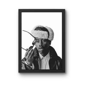 Puede incluir: Retrato enmarcado en blanco y negro de una persona con sombrero de piel, gafas de sol, camisa a rayas y chaqueta de cuero. La persona sostiene las gafas de sol y mira al espectador. La imagen es un retrato de estudio.