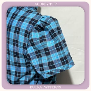 May include: A short-sleeved top labeled AUDREY TOP by BUGRA PATTERNS, featuring a bright blue and black plaid pattern. The garment has gathered puff sleeves with a straight hem and a structured shoulder design, showcasing a woven fabric texture.