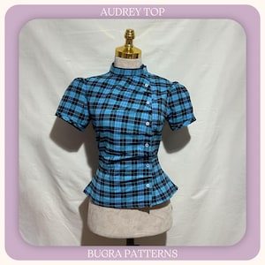 May include: A short-sleeved top with a blue and black plaid pattern, featuring a high collar and an asymmetrical button-down front closure. The garment is displayed on a mannequin and includes the text AUDREY TOP and BUGRA PATTERNS.