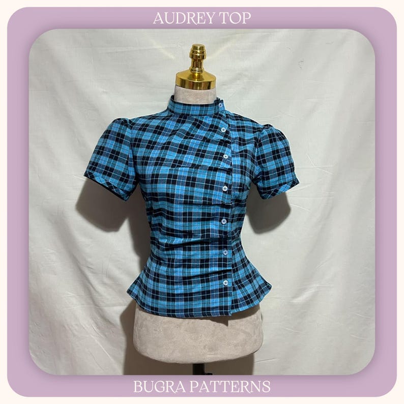 May include: A short-sleeved top with a blue and black plaid pattern, featuring a high collar and an asymmetrical button-down front closure. The garment is displayed on a mannequin and includes the text AUDREY TOP and BUGRA PATTERNS.