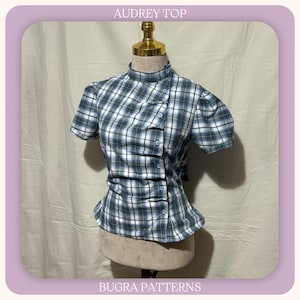 May include: A short-sleeved top with a blue, white, and green plaid pattern, featuring a high collar and an asymmetrical button-down front closure. The garment is displayed on a mannequin and includes the text AUDREY TOP and BUGRA PATTERNS.