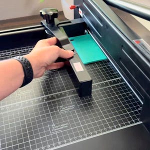 Może przedstawiać: Czarna gilotyna do papieru z podstawą w kratkę, białymi liniami pomiarowymi oraz oznaczeniami format&oacute;w papieru A4 i B5. Urządzenie posiada regulowane czarne ramię prowadzące z pokrętłem oraz metalowe ostrze tnące z czerwonymi akcentami.
