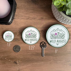 three circular badges with the text "WILD AUGUST" printed on them, placed on a wooden surface alongside a key and a plant.