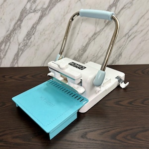 May include: A white and light blue Cinch book binding machine featuring a metal handle, a white base with a black logo, and a light blue platform with the text Wild August embossed on the surface. The device includes a punch mechanism.