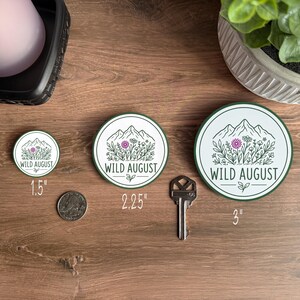 three circular badges with the text "WILD AUGUST" printed on them, placed on a wooden surface alongside a key and a plant.
