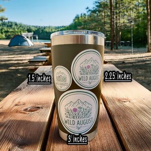 A 16oz tumbler with three stickers on it, placed on a wooden picnic table in a forested area.