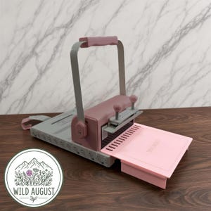 May include: A dusty rose and light gray paper binding machine featuring a vertical handle, a punch mechanism with two knobs, and a flat base. A light pink plastic cover with a scalloped edge and the text WILD AUGUST is attached to the front.