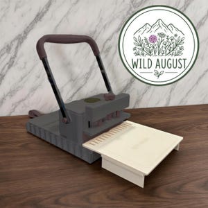 May include: A dark gray bookbinding punch tool with a tall handle and a cream-colored base plate featuring a series of rectangular slots. A circular logo in the upper right corner displays a mountain illustration and the text WILD AUGUST.