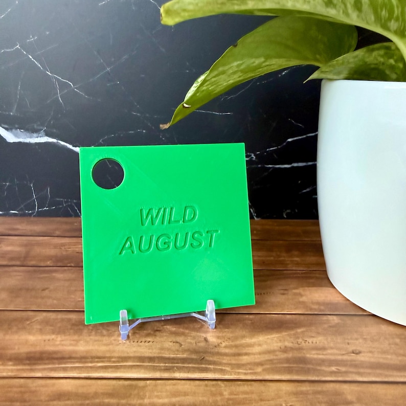 Może przedstawiać: Kwadratowa, jasnozielona plastikowa zawieszka z wytłoczonym napisem WILD AUGUST na środku oraz okrągłym otworem w lewym g&oacute;rnym rogu. Zawieszka jest umieszczona na małej, przezroczystej plastikowej podstawce o gładkiej powierzchni.