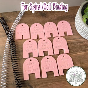 May include: A set of nine pink arch-shaped plastic spiral coil binding size guides, each engraved with metric and imperial measurements ranging from 6mm (1/4 inch) to 25mm (1 inch). Two metal spiral coils in black and white are positioned to the left.