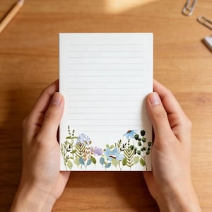 May include: A lined notepad with a floral design. The notepad is held by hands and features a watercolor illustration of blue, purple, and green flowers along the bottom edge. The background is a wooden surface.