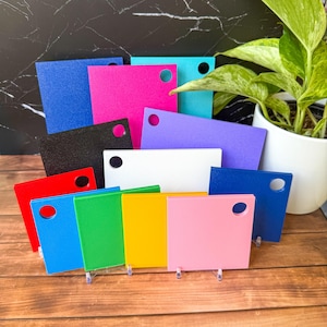 Peut inclure: Une collection d'échantillons rectangulaires en acrylique de différentes couleurs unies, notamment bleu, rose, noir, violet, blanc, rouge, vert, jaune et rose clair. Chaque échantillon présente un trou circulaire percé près d'un coin supérieur.