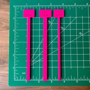 May include: Three bright pink T-shaped measuring tools with labels. The first reads 1/4 inch and 6mm, the second 5/16 inch and 8mm, and the third 5/32 inch and 4mm. These tools are positioned on a green cutting mat with white grid lines.