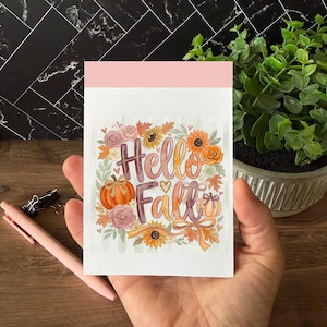 May include: A rectangular notepad with a light pink top border and a white background featuring a watercolor illustration of pumpkins, sunflowers, roses, and autumn leaves surrounding the text Hello Fall in orange and purple script lettering.