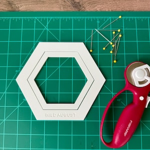 Op de afbeelding: Een wit zeshoekig quilt-sjabloon met de tekst WILD AUGUST in reliëf op de onderrand, geplaatst op een groene snijmat naast een rode rolsnijder en verschillende spelden met gele kopjes verspreid over het oppervlak.