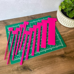 May include: A set of nine bright pink acrylic seam allowance guides of varying widths, each featuring engraved text indicating measurements in both inches and millimetres, arranged in a row on a green self-healing cutting mat.