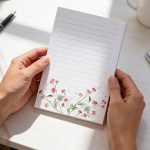 May include: A lined notepad with a floral design. The notepad is held by a person. The bottom of the notepad features a watercolor design of pink flowers and green leaves with a speckled background. The notepad is white with gray lines.