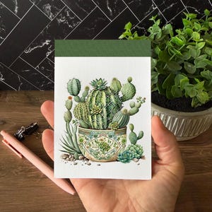 May include: A rectangular notepad with a green top border and a white background featuring a detailed illustration of various green cacti planted in a decorative, patterned ceramic pot with blue, green, and gold geometric and floral designs.