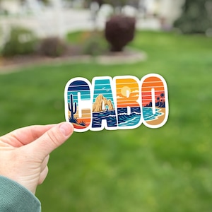 May include: A die-cut vinyl sticker featuring the word CABO in bold block letters. The design incorporates a colorful landscape scene with a cactus, a coastal rock arch, a sunset, ocean waves, and palm trees in shades of blue, orange, and yellow.