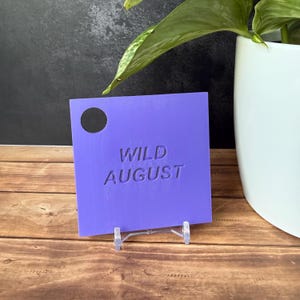 Może przedstawiać: Kwadratowa fioletowa zawieszka z wytłoczonym napisem WILD AUGUST na środku oraz okrągłym otworem w lewym g&oacute;rnym rogu. Zawieszka umieszczona jest na małym, przezroczystym plastikowym stojaku i ma gładkie, matowe wykończenie.