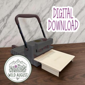 Op de afbeelding: Een grijs papierponsapparaat met een donker handvat en een crèmekleurige basisplaat. De tekst DIGITAL DOWNLOAD wordt weergegeven in paars schrift. Een rond logo toont een bergketen, bloemen en de tekst WILD AUGUST in zwarte inkt.
