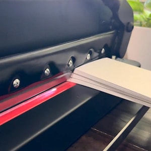Może przedstawiać: Czarna gilotyna do papieru z ostrą metalową głowicą i czerwoną osłoną bezpieczeństwa. Stos białego papieru jest umieszczony pod ostrzem, gotowy do cięcia. Urządzenie posiada solidną metalową ramę z widocznymi srebrnymi śrubami wzdłuż g&oacute;rnej części.