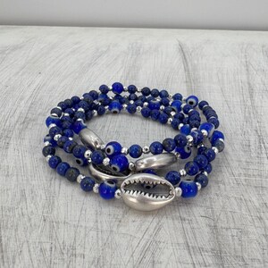 May include: A layered bracelet featuring dark blue beads, silver accents, and eye-shaped beads. The bracelet also includes a silver seashell charm. The jewelry is displayed on a light gray, textured surface.