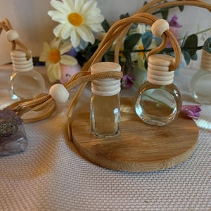 Könnte beinhalten: Klare Aromatherapie-Flaschen aus Glas mit Holzdeckeln und beigefarbenen Kordeln. Die Flaschen stehen auf einer runden Holzbasis. Ein violetter Kristall und Blumen sind im Hintergrund. Die Flaschen sind mit einer klaren Flüssigkeit gefüllt.